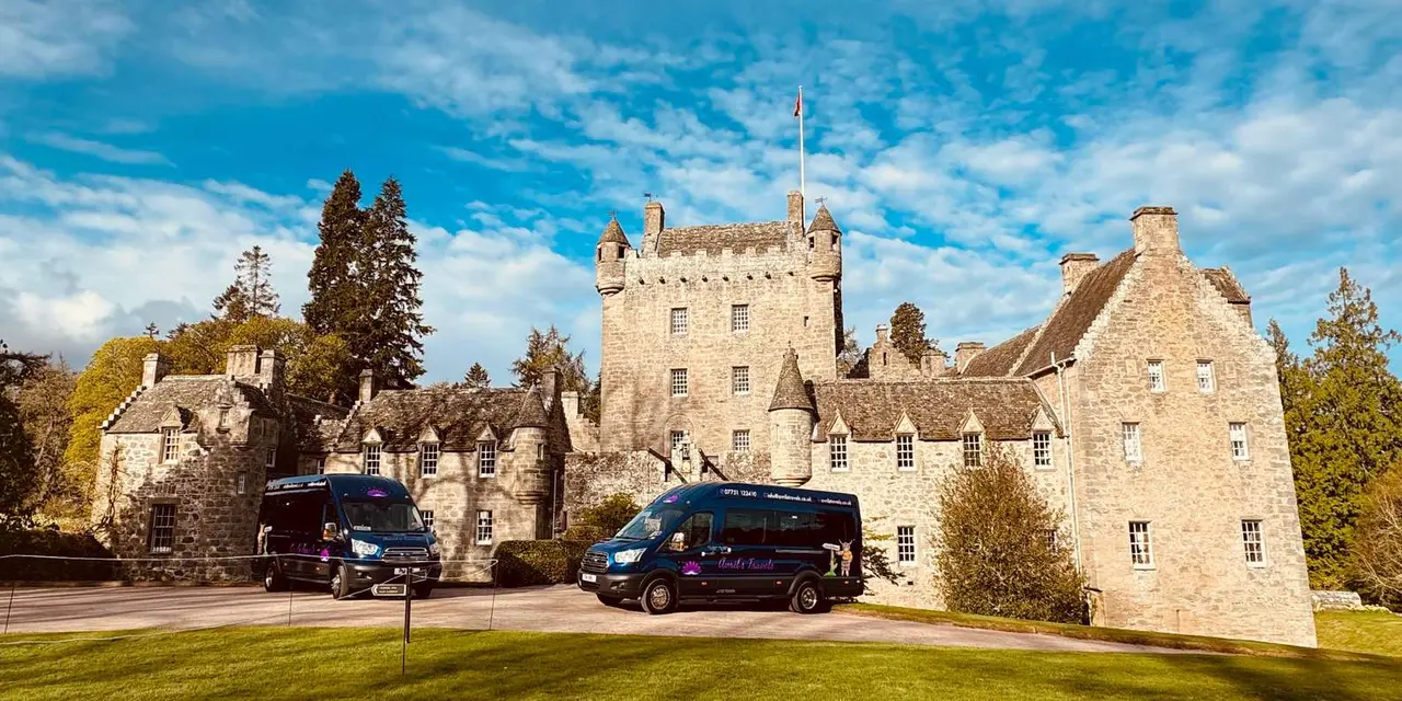 Two navy minibuses parked on the gravel in front of a pale stone castle under a blue sky