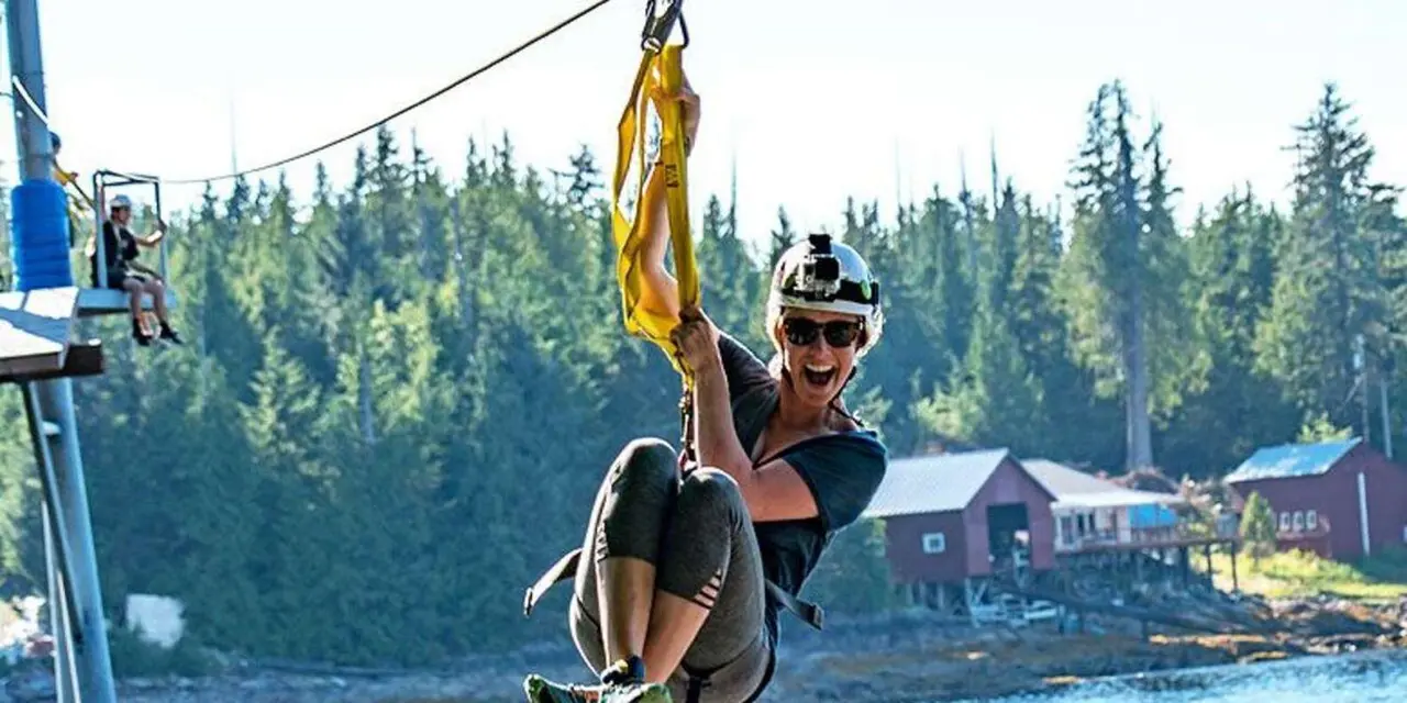 A rider on a zipline above the water with forest and buildings behind