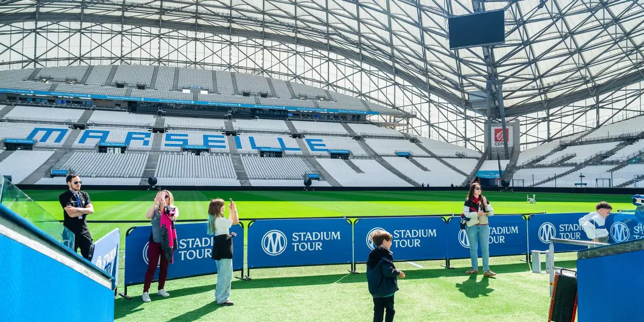 Visitors walking on the track beside the pitch in front of Stadium Tour boards, under the Vélodrome's curved roof