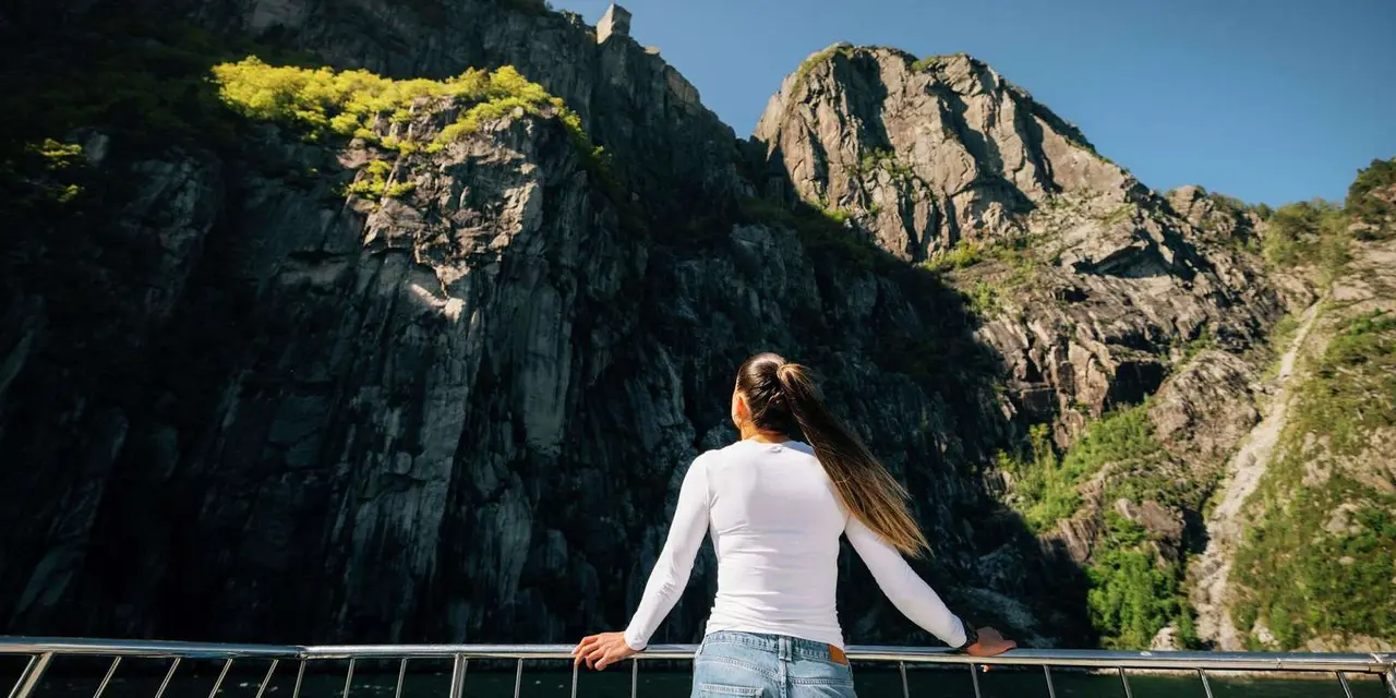A woman in a white top at a boat rail looking up at sunlit fjord cliffs