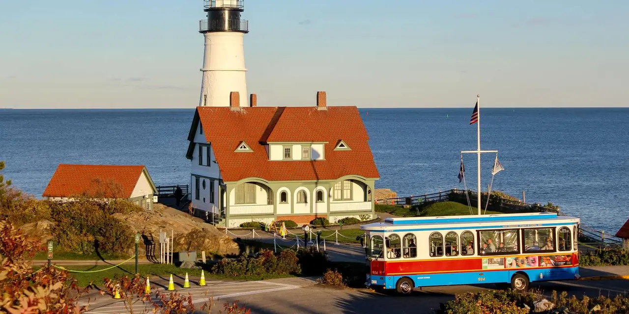 A red, white and blue trolley parked beside Portland Head Light, a white lighthouse with a red-roofed keeper's house, on a sunny day