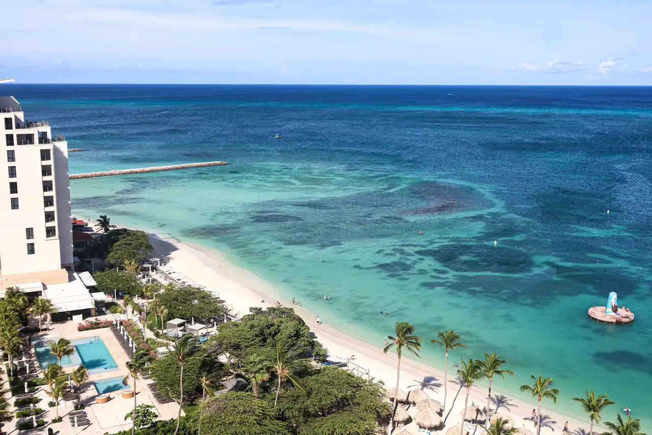 A curve of white-sand beach on Aruba's north-west coast, with turquoise shallows fading to deep blue, palm trees, thatched beach umbrellas and a resort tower seen from above