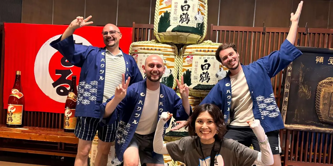 A cheerful tour group in blue happi coats posing with a guide in front of a sake brewery display