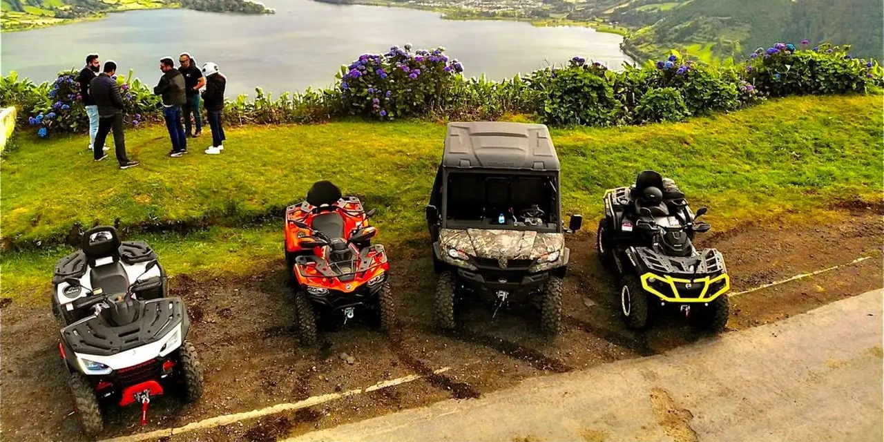 Quad bikes and a buggy parked on a grassy rise above a crater lake, riders by a hydrangea hedge