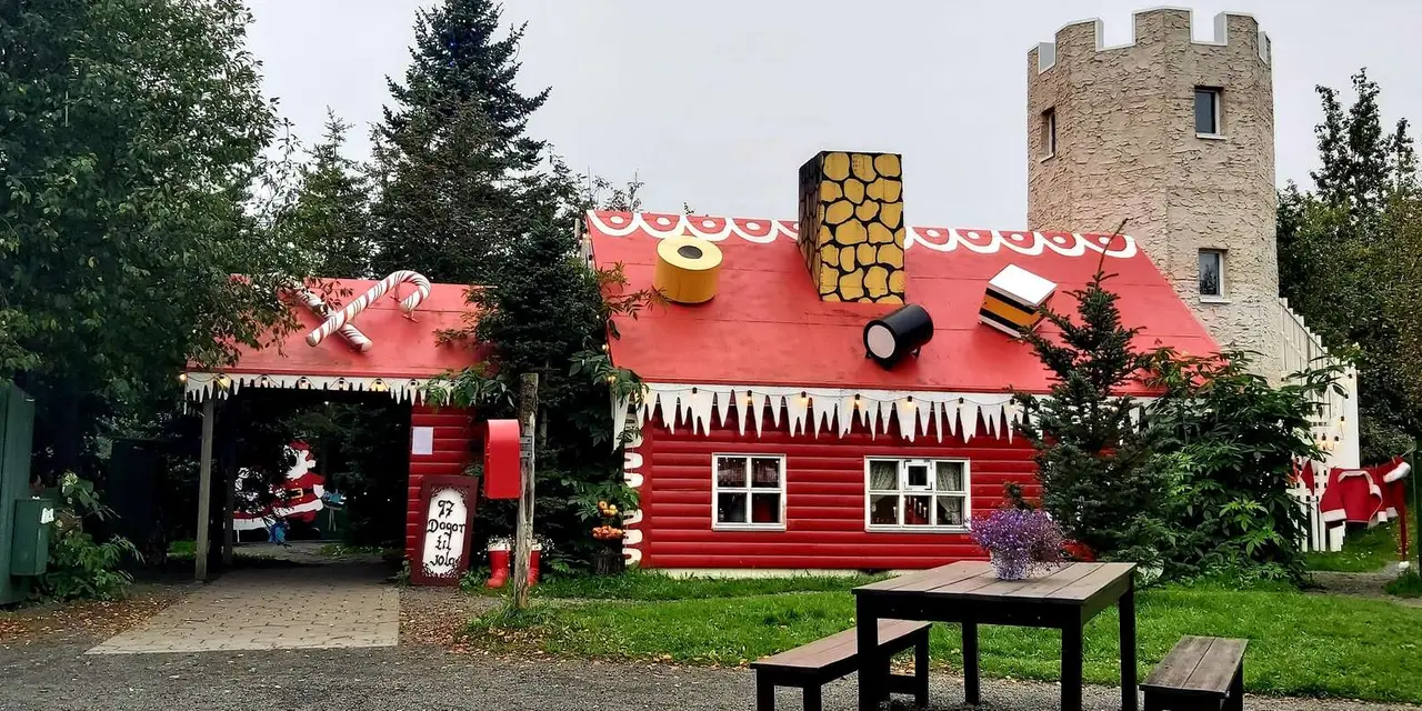A red log-style Christmas House with white icicle trim and a Santa figure in the doorway beside a picnic table