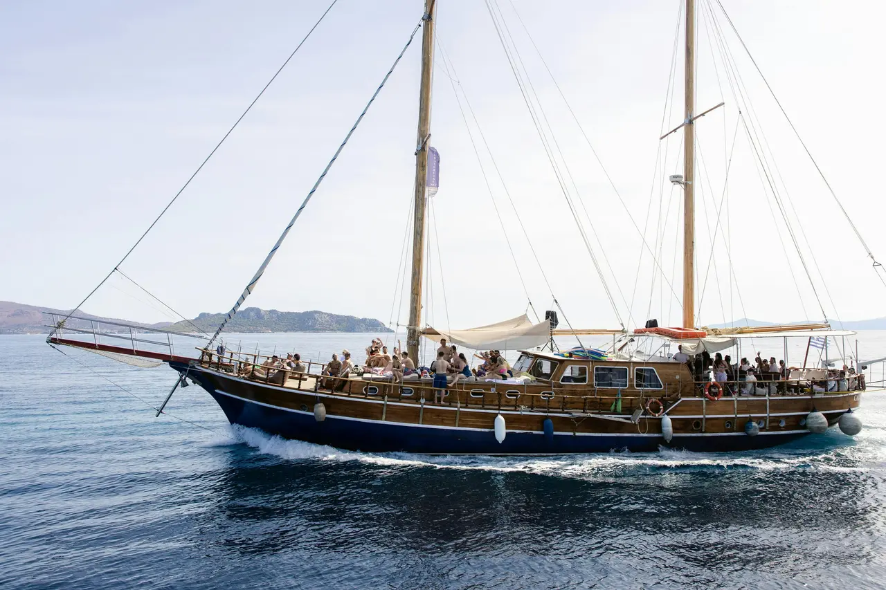 A two-masted wooden sailing boat cruising on calm blue water with passengers relaxing on deck