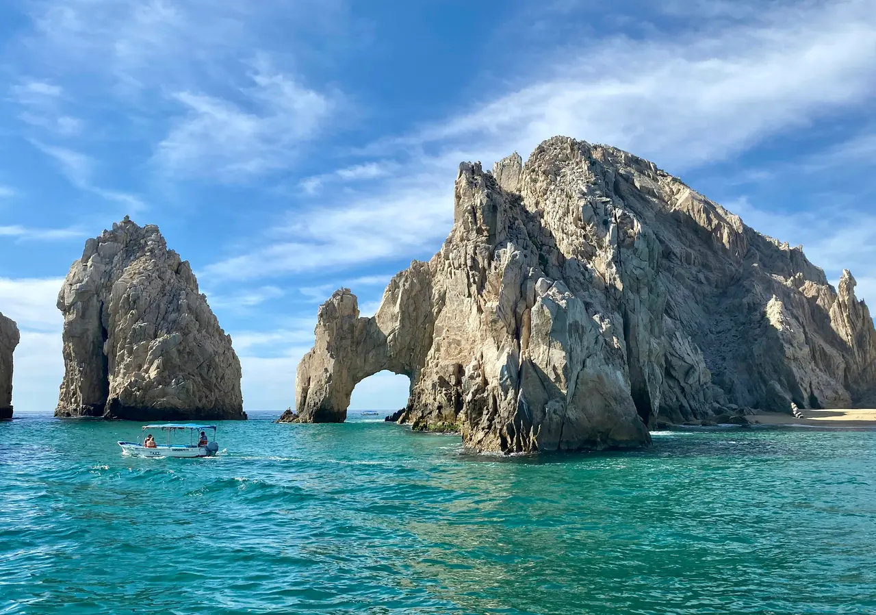 The granite Arch at Land's End in Cabo San Lucas, with a small tour boat on turquoise water and a streaky blue sky