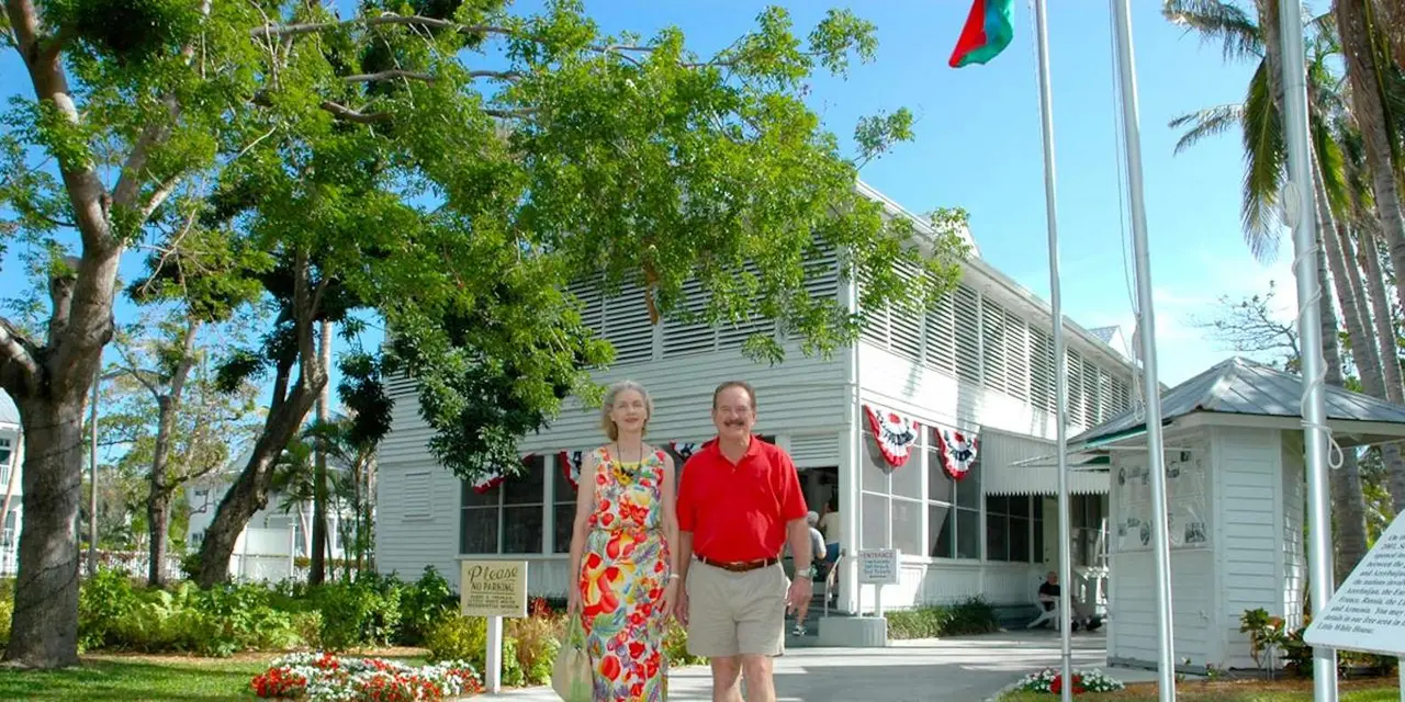 Two visitors walking out of the white, wood-slatted Truman Little White House past bunting, palms and a flagpole