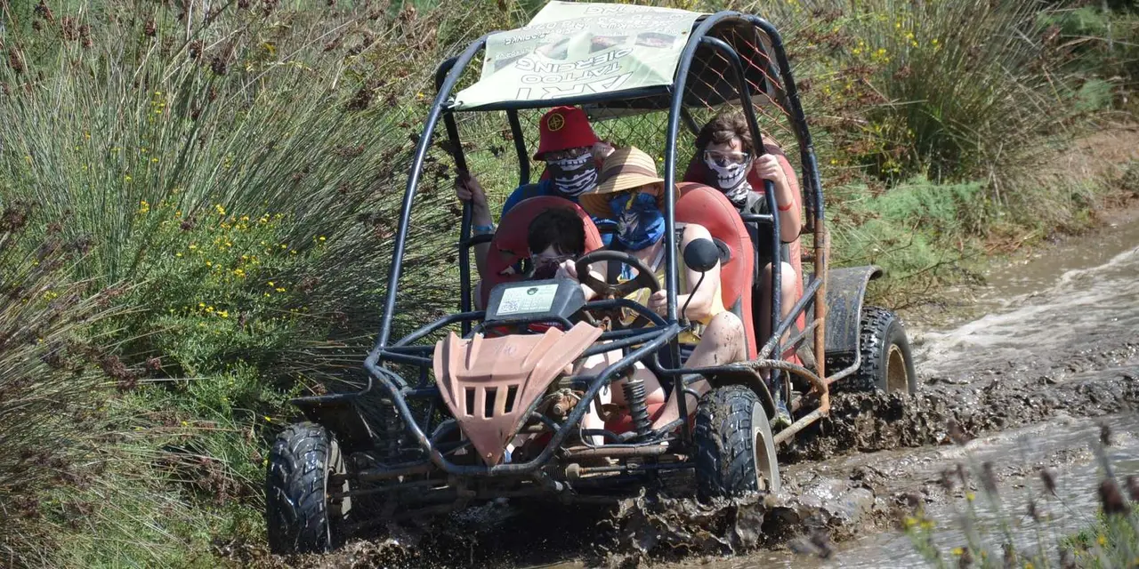 A buggy with two riders driving through a muddy puddle on a wooded dirt trail