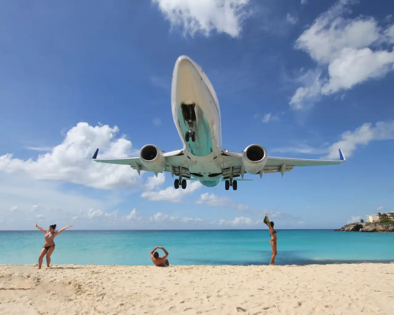 A passenger jet descending low over a white-sand beach and turquoise water at Maho Beach in Sint Maarten, with three sunbathers looking up