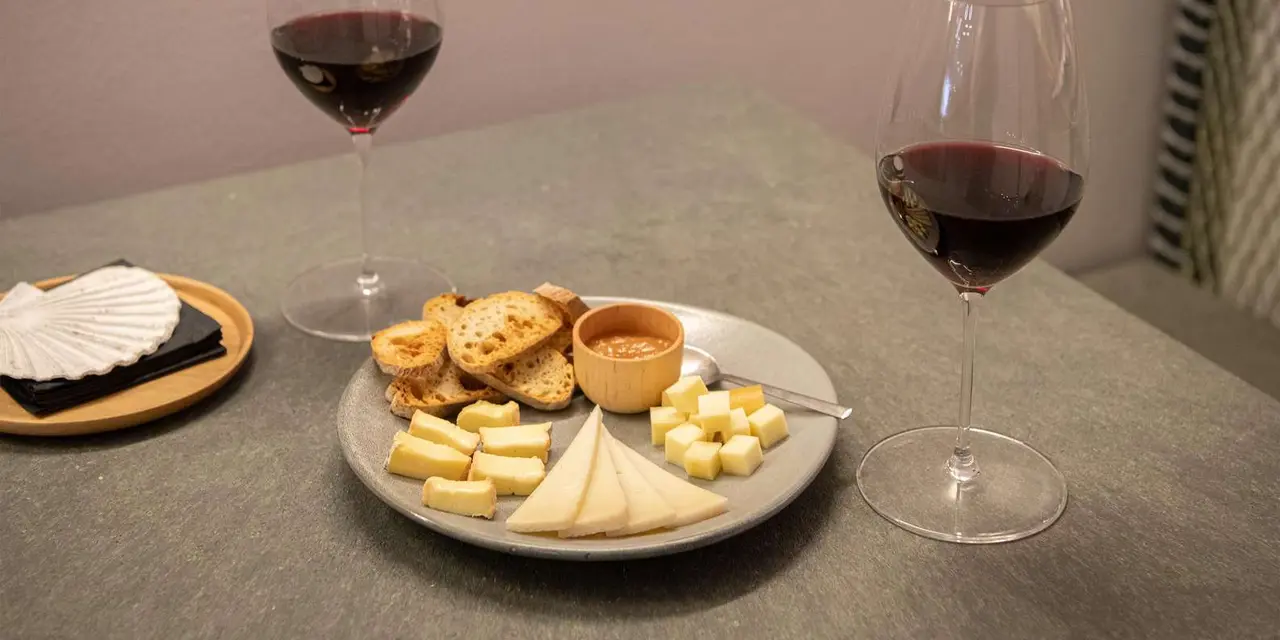 A glass of red wine beside small plates of cheese cubes and bread on a table