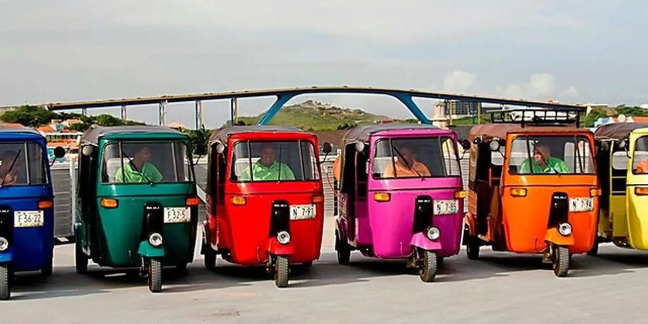 Four brightly painted tuk-tuks in green, red, pink and orange lined up beneath a high bridge