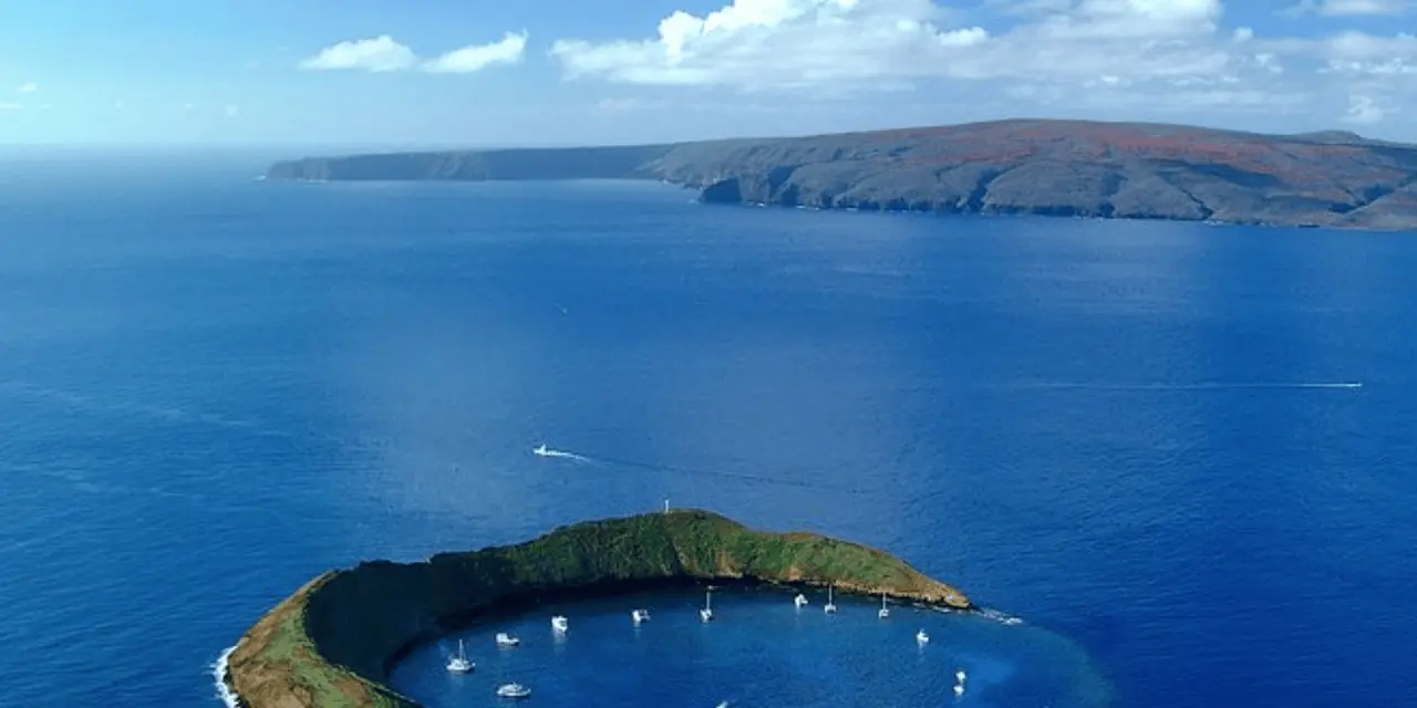 An aerial view of Molokini, a crescent-shaped islet with a handful of boats moored inside its curve, and a long dark island beyond