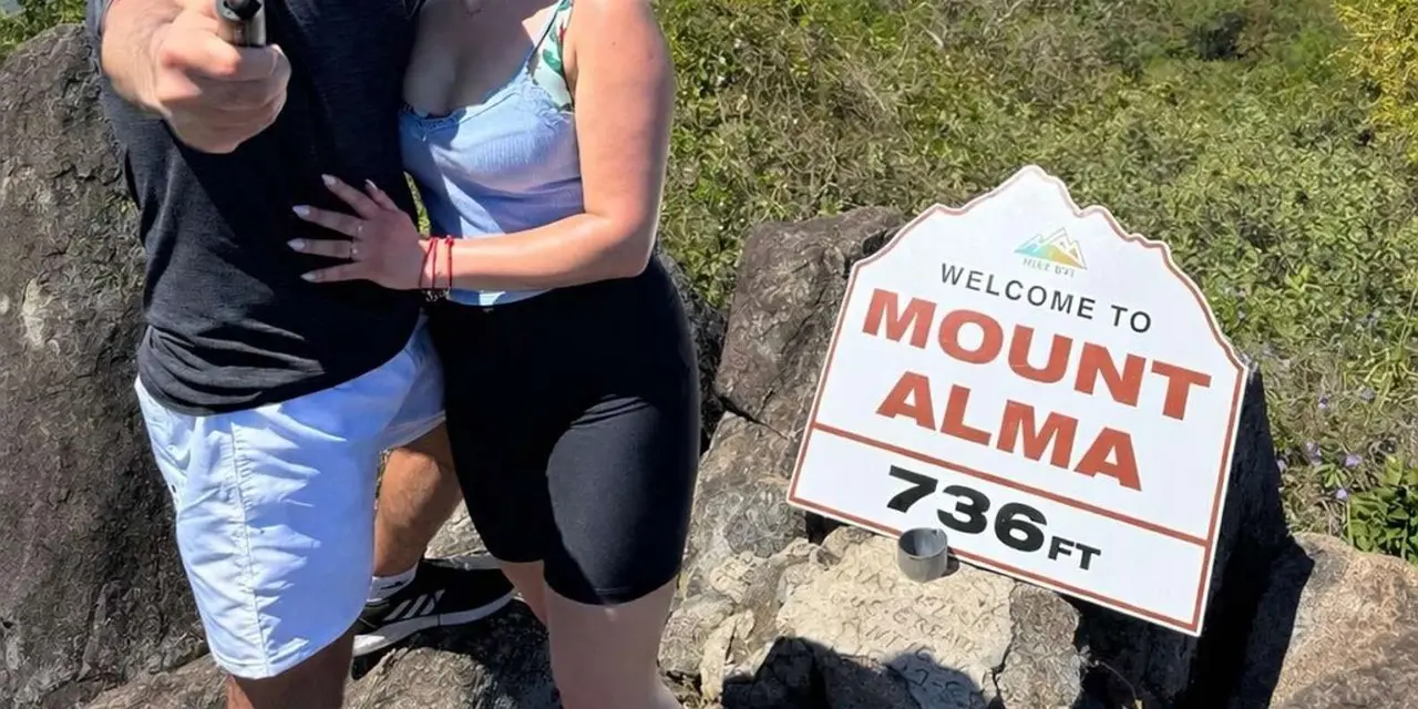 A couple taking a selfie beside a 'Welcome to Mount Alma 736 ft' sign, with bays and islands far below