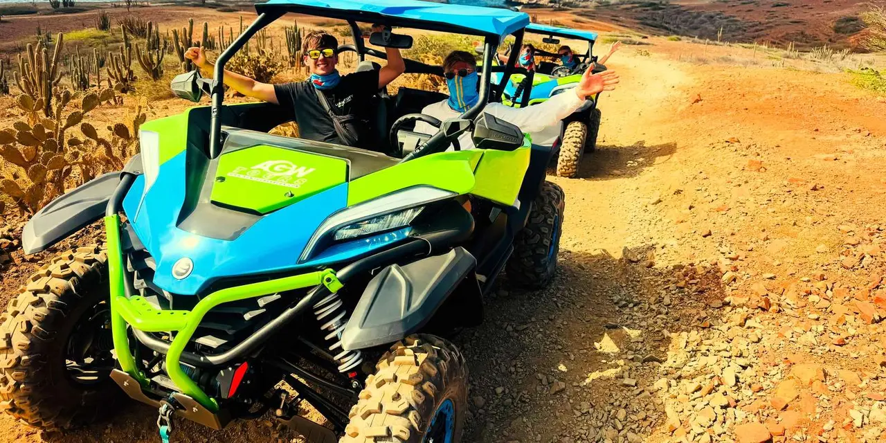 Two blue-and-green UTVs driving in line along a red-dirt track between cacti, the driver in front waving