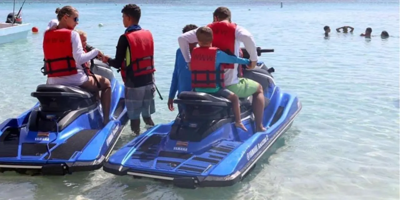 People in red life vests on two blue jet skis in shallow turquoise water, with a ship far out on the horizon