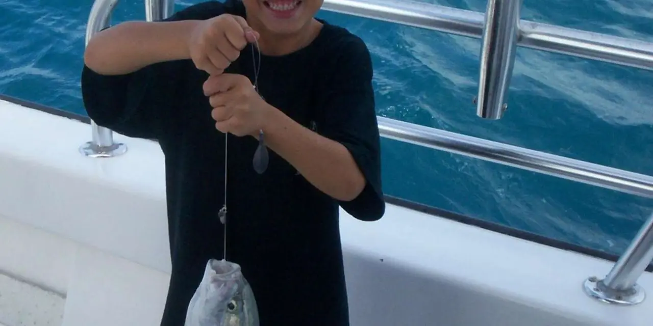 A young boy on a boat deck holding up a small fish he caught