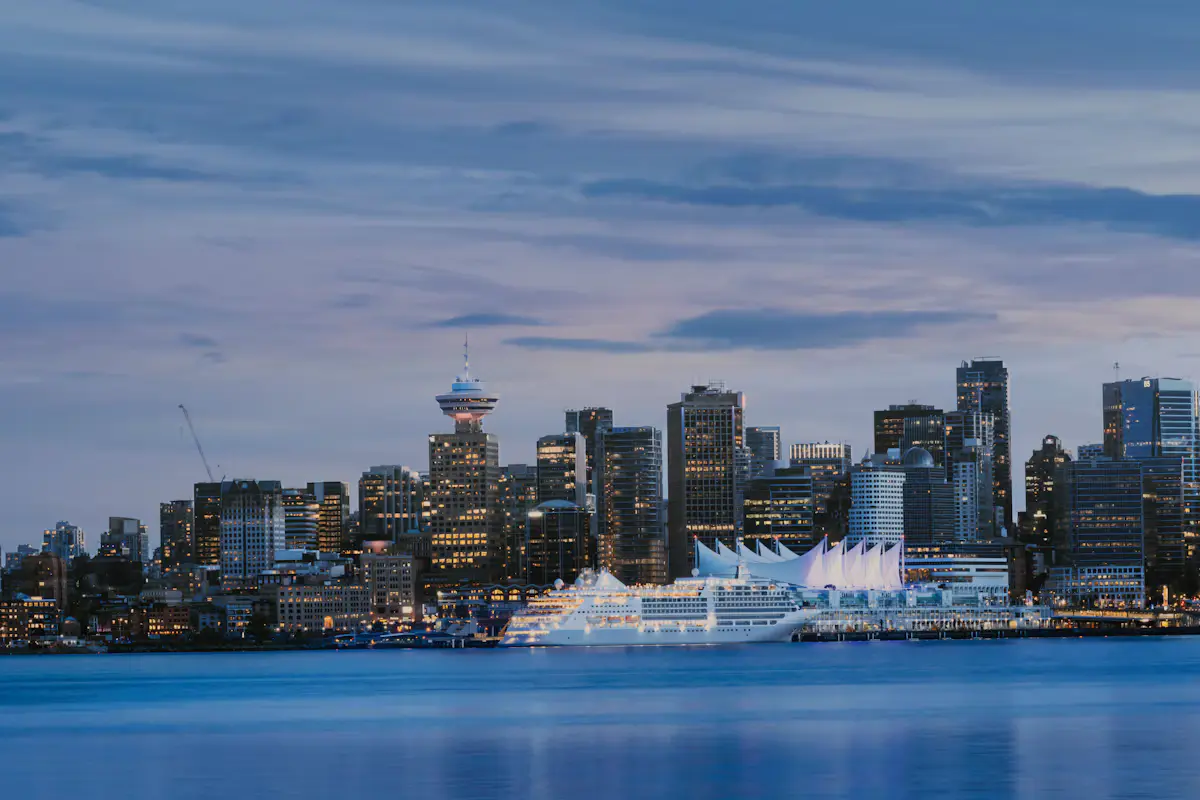 Vancouver's waterfront at dusk seen across the harbour: a lit cruise ship docked beside the white sails of Canada Place, with downtown towers behind under a blue evening sky