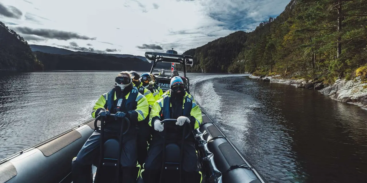 Passengers in yellow-and-black flotation suits seated on a RIB speeding across a fjord under cloudy skies