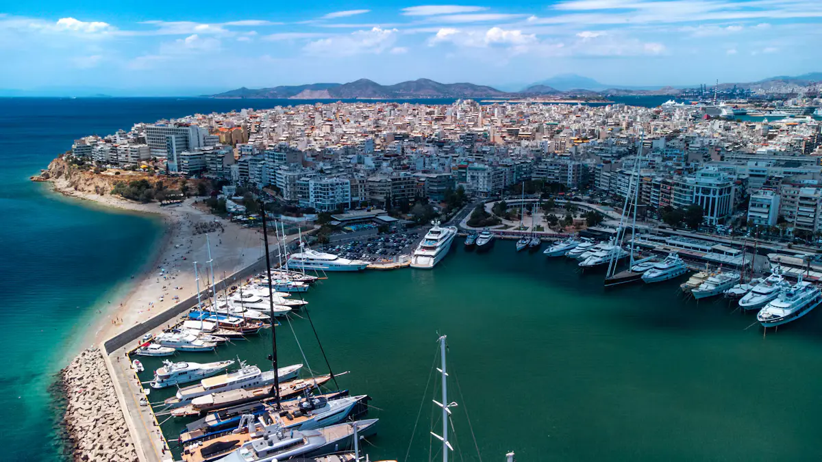 Aerial view of a Piraeus marina lined with white yachts, a narrow sandy beach along the breakwater and pale apartment blocks climbing the hill behind