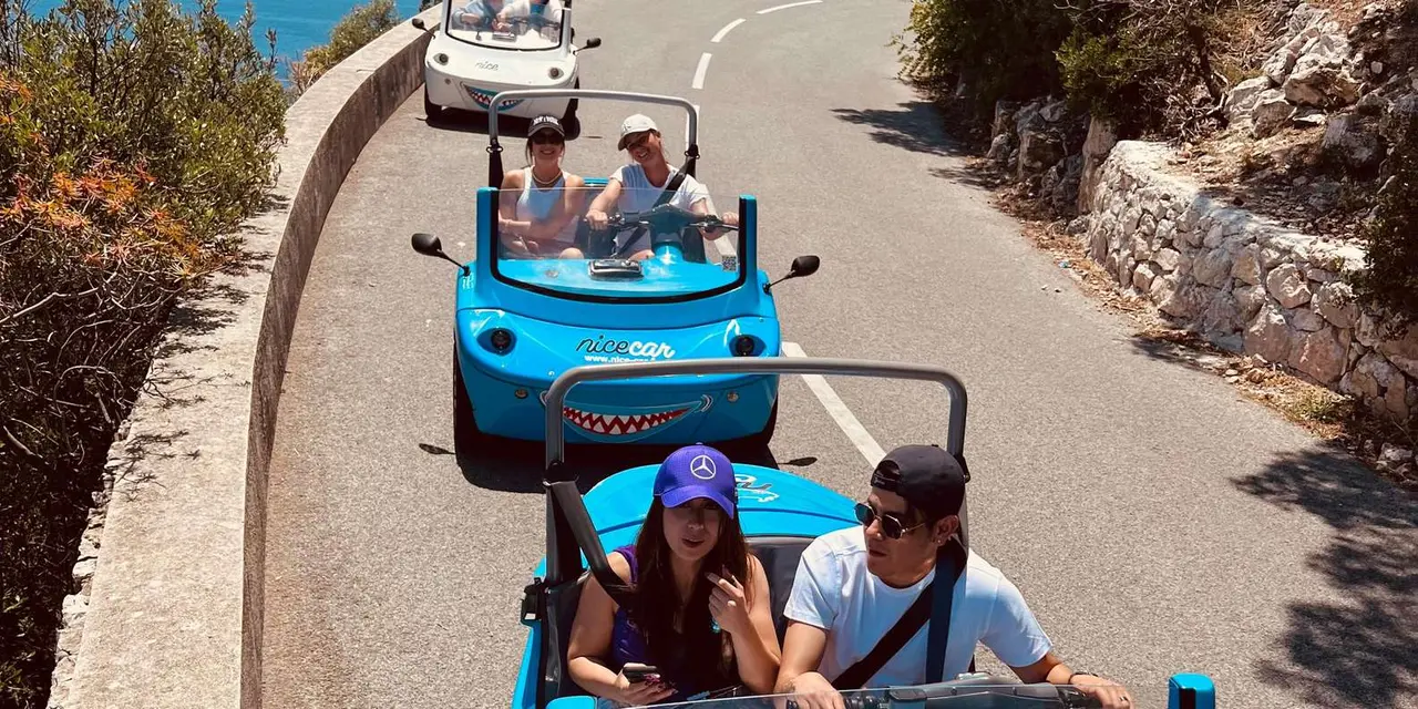 A line of small blue open-top electric cars driving a winding coastal road above the sea