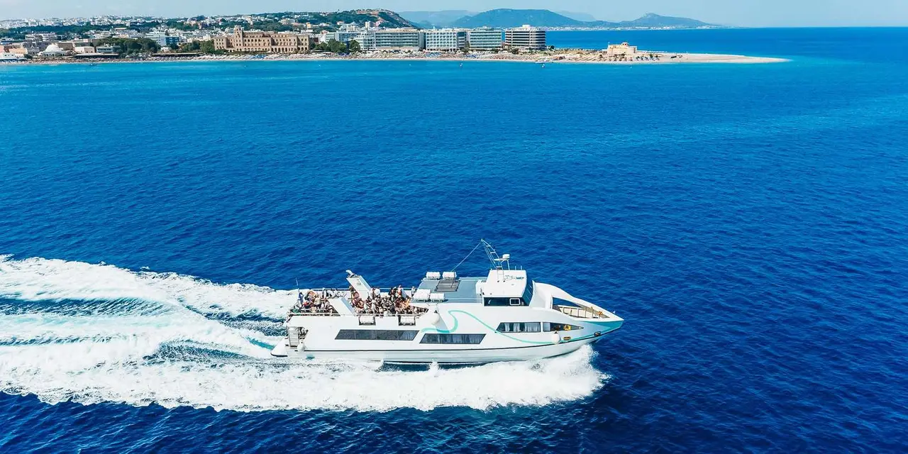 A white speedboat leaving a long wake across deep blue water, with the Rhodes shoreline and hotels behind