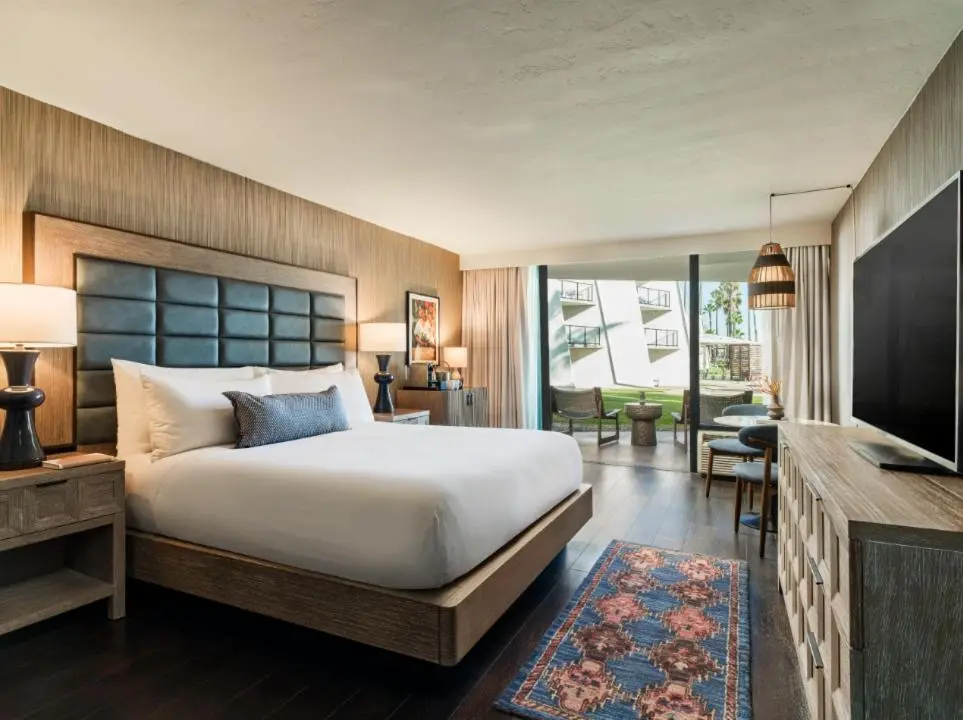 A king bedroom at The Maya in Long Beach, with a padded headboard, a patterned rug and glass doors out to a courtyard lawn