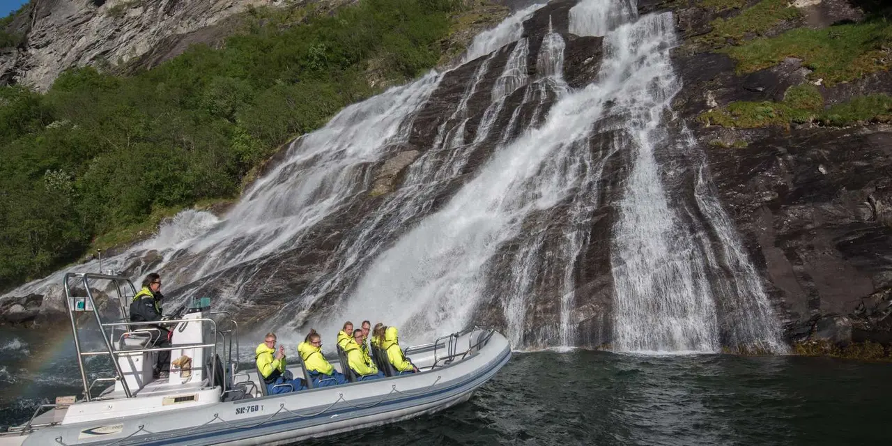 A grey RIB boat full of passengers in yellow suits at the foot of a broad waterfall pouring into the fjord
