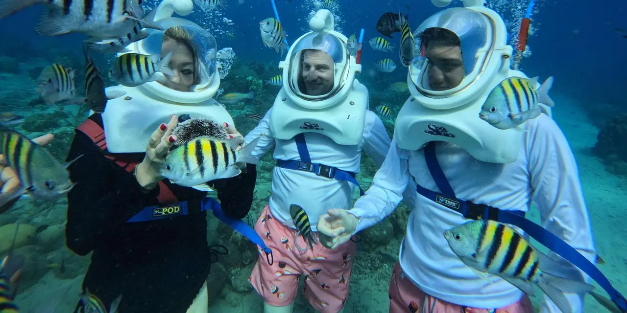 Three people in large diving helmets standing on the seabed, surrounded by striped sergeant major fish