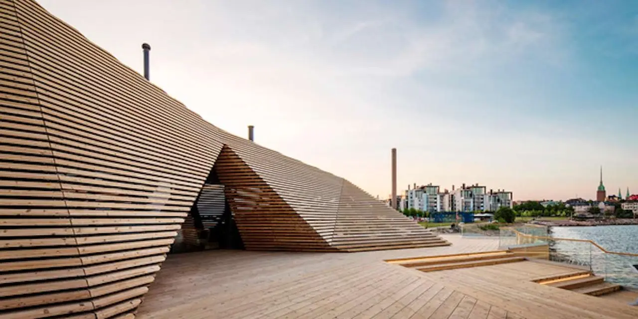 The angular wooden Löyly sauna building at the water's edge, with a boardwalk in front and apartment blocks on the far shore under a pale evening sky