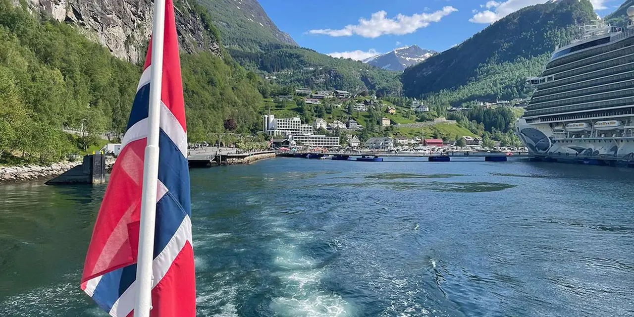 A Norwegian flag flying at the stern of a boat, with Hellesylt village and mountains behind