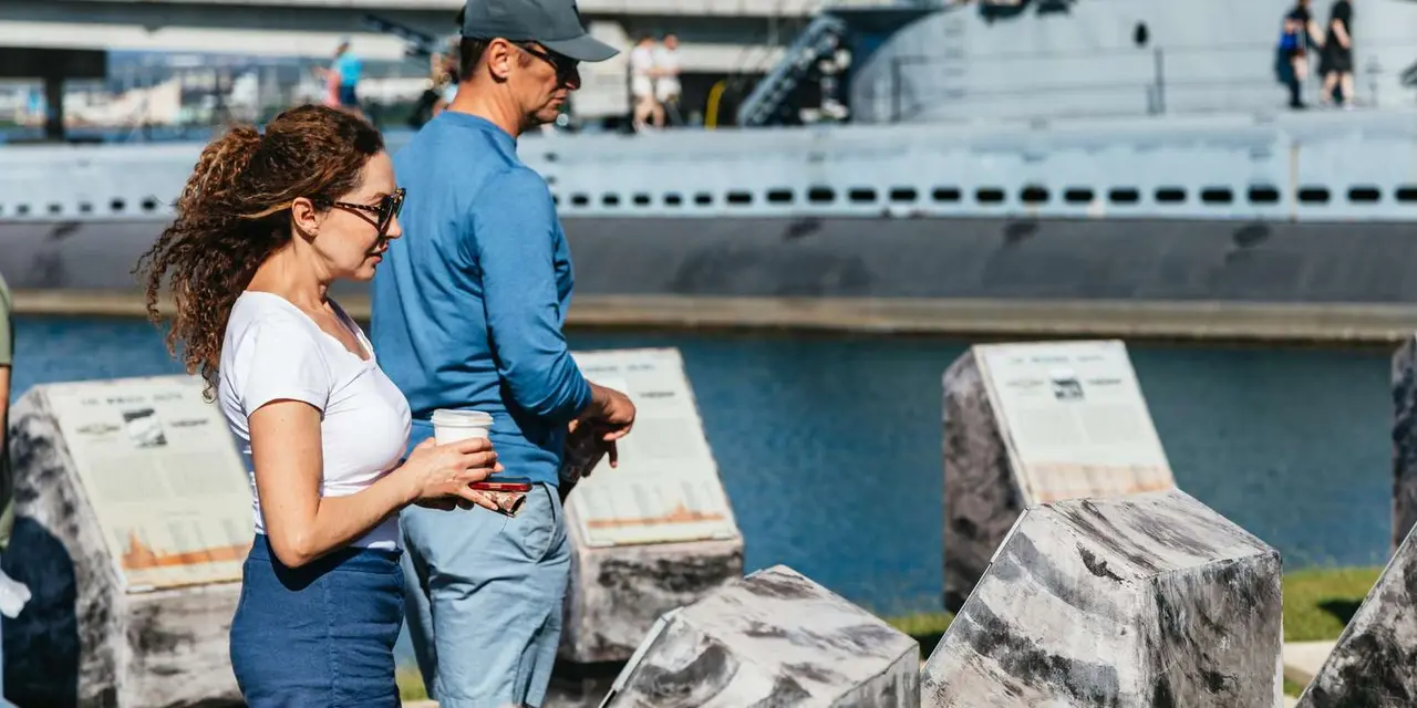 A couple reading an exhibit on the Pearl Harbor waterfront, with a grey warship behind them
