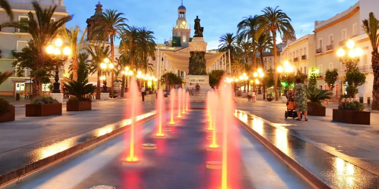 Plaza de San Juan de Dios in Cádiz at dusk, with lit fountains in the foreground, palm trees and the illuminated Town Hall clock tower behind