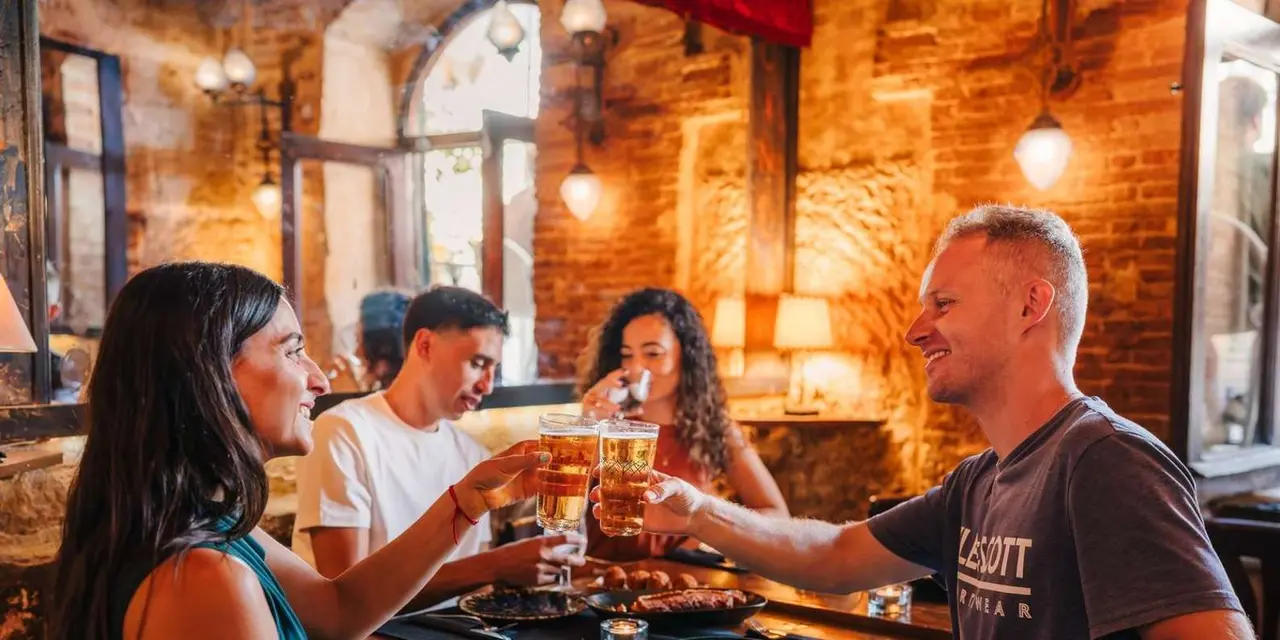 Four people raising beer glasses over a table of food in a warmly lit stone-walled tavern