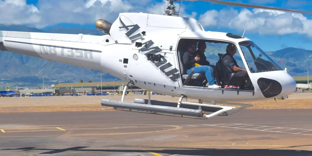 A white Air Maui helicopter lifting off the heliport apron with a pilot and two passengers visible through the open door, mountains and airport buildings behind