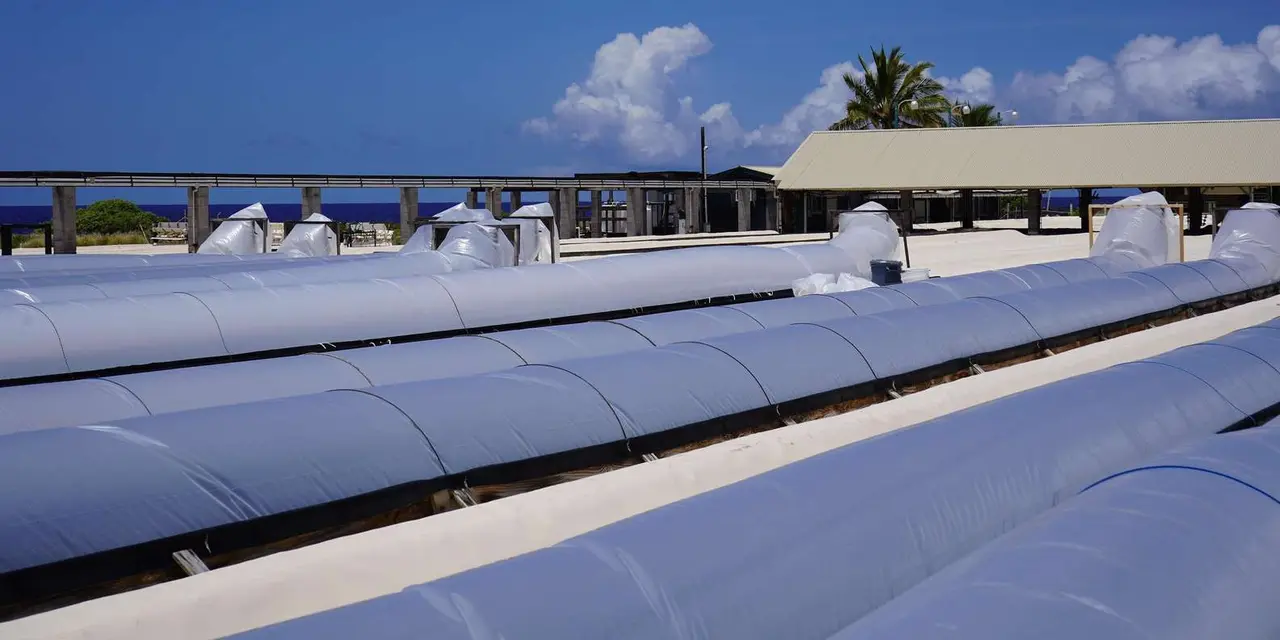 Rows of long covered salt-drying troughs on an oceanfront farm under a blue sky