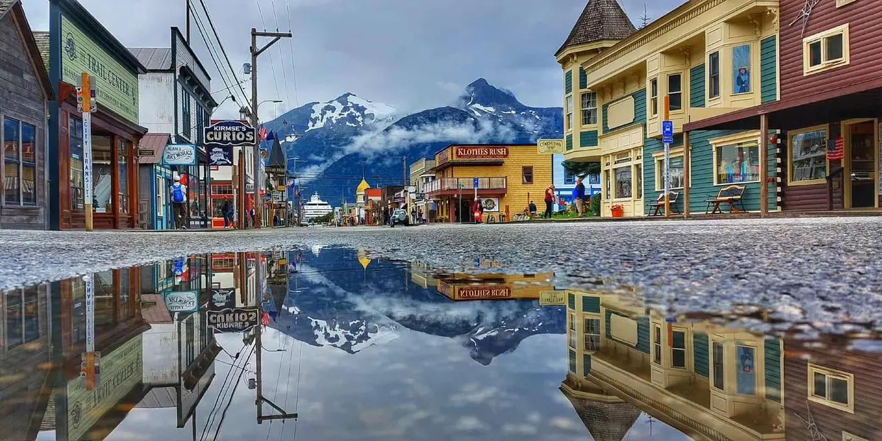 Skagway's main street with snowy mountain peaks above it, mirrored in a puddle