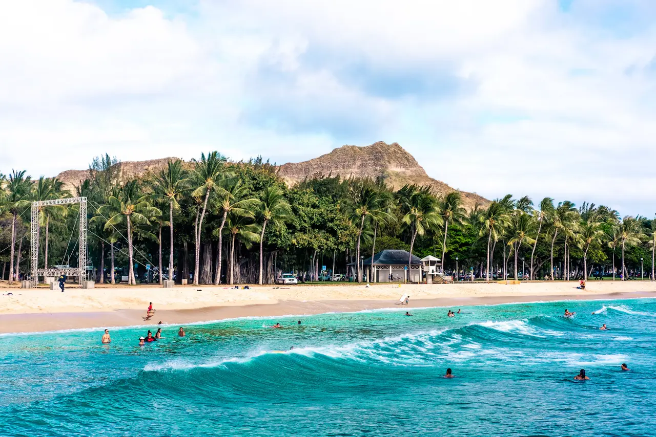 Swimmers and boogie boarders in turquoise surf off Kūhiō Beach in Waikīkī, with palm trees and Diamond Head behind; Waikīkī is about 3 miles from the cruise piers