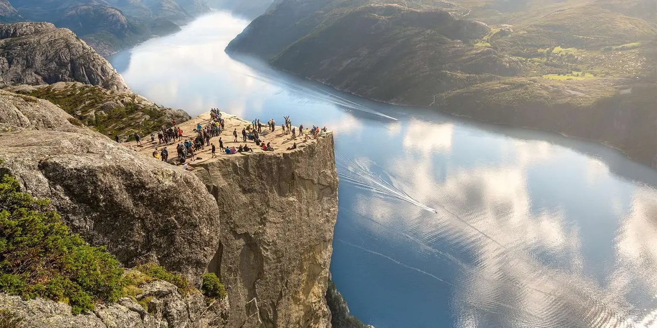 People standing on the edge of Pulpit Rock above a still fjord that mirrors the sky