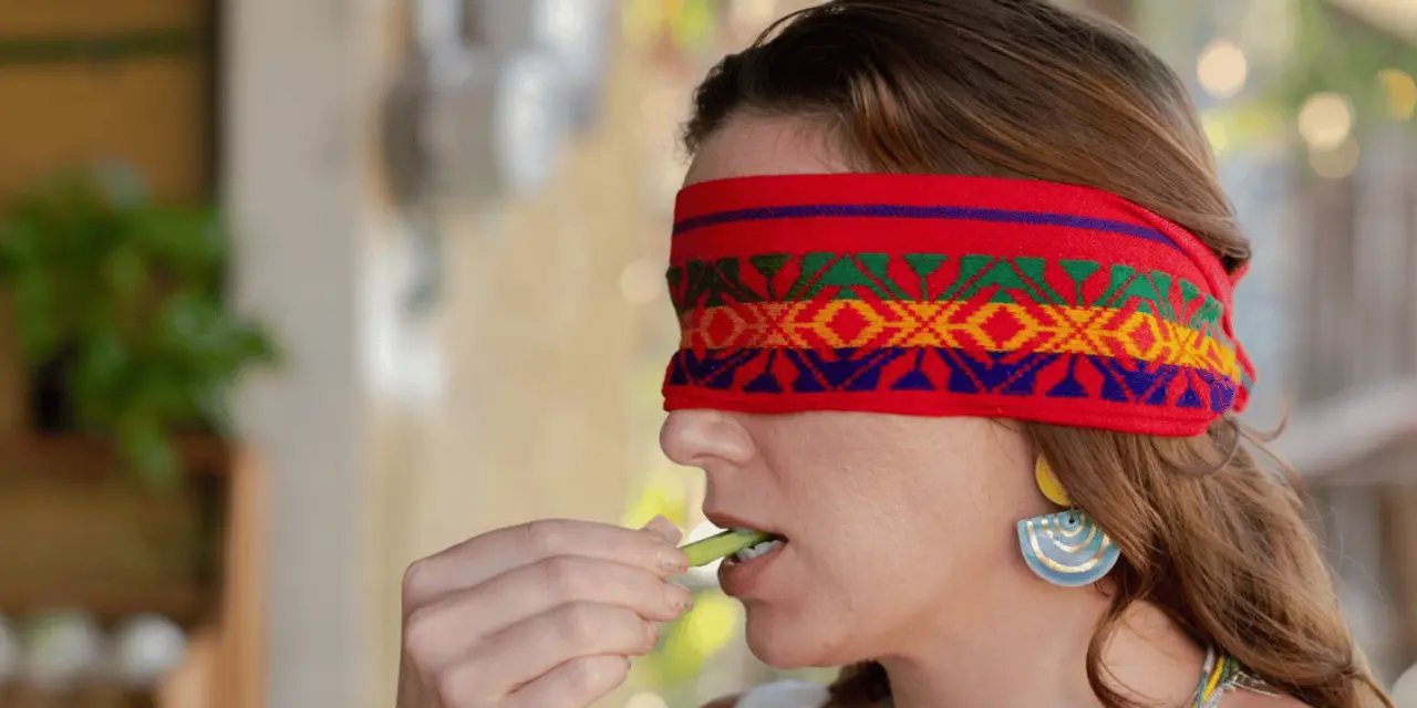 A woman wearing a red patterned blindfold about to taste a small green item held in her fingers