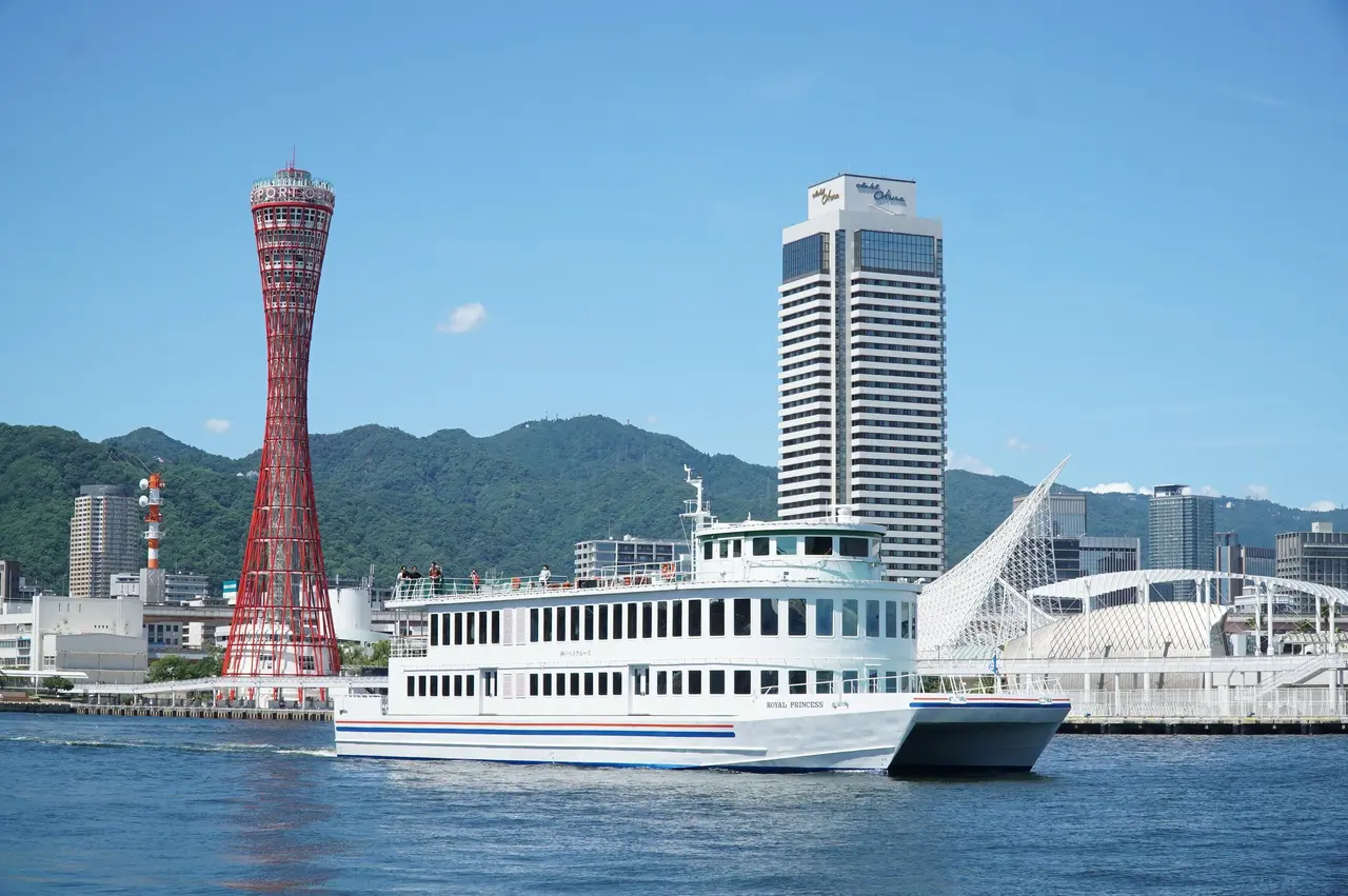 A white sightseeing boat in Kobe harbor in front of the red Port Tower and the Hotel Okura on a clear day