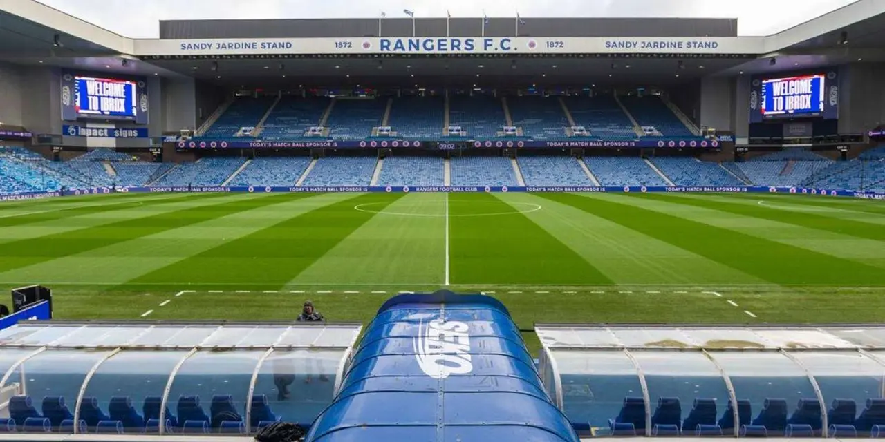 Ibrox Stadium's empty stands and striped pitch, seen from behind a blue dugout roof with the Rangers crest
