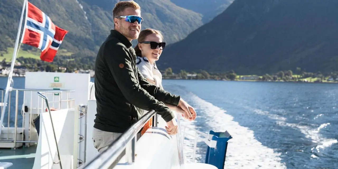 A couple at the rail of a boat on a fjord beside a Norwegian flag, with mountains behind