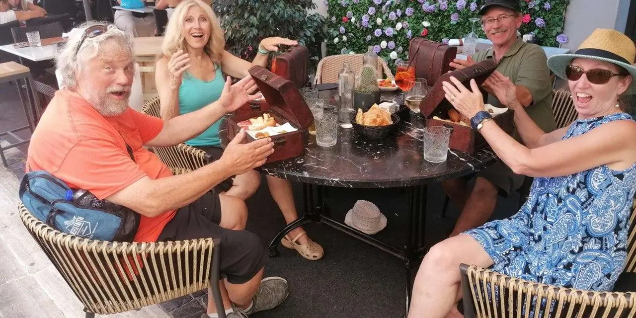 Four people laughing around a cafรฉ table covered with plates of food, with a wall of flowers behind them