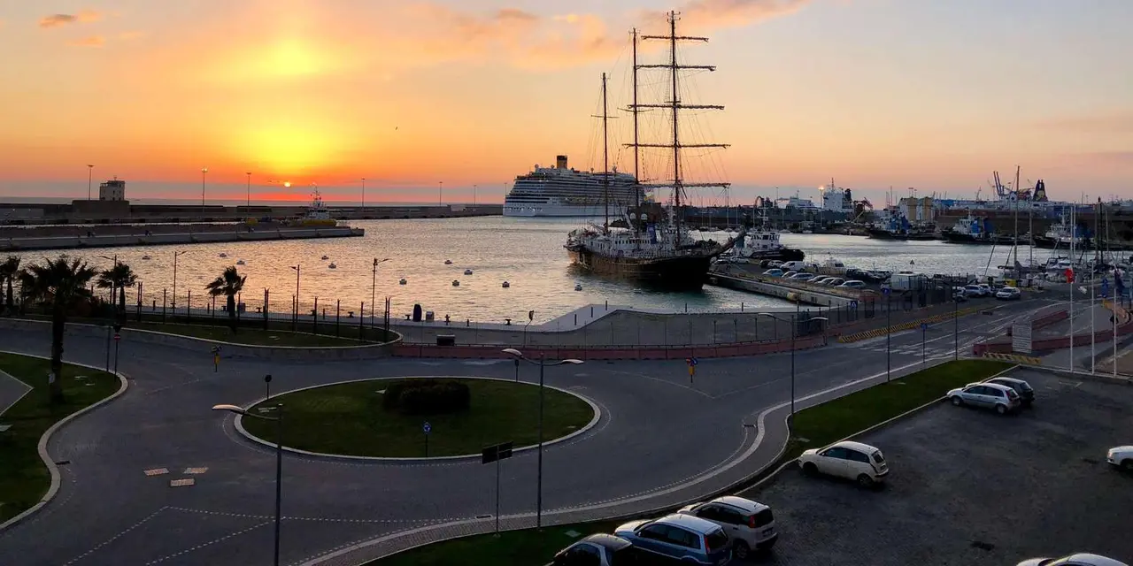 Sunset over Civitavecchia harbor, with a tall sailing ship and a cruise ship moored beyond a waterfront roundabout