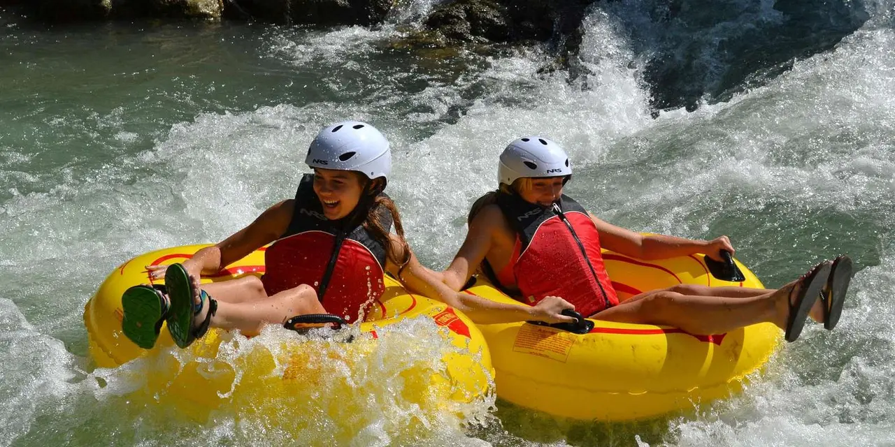 Two people in helmets and life vests riding yellow inner tubes through white water