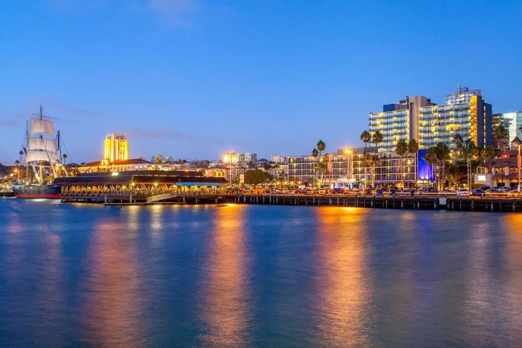 The Wyndham San Diego Bayside's lit-up towers on the waterfront at dusk, across blue harbor water with a tall ship moored at the left