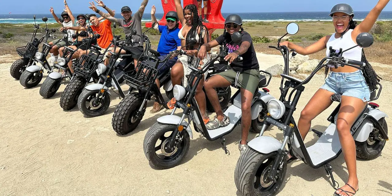 A group of riders in helmets on a row of fat-tyre scooters on sand beside the sea, arms in the air