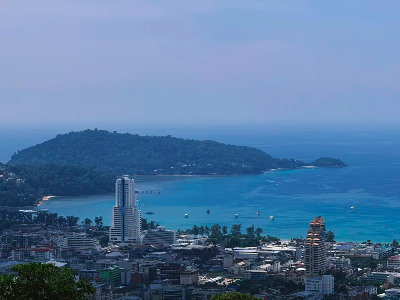 View over Patong's rooftops and towers to the turquoise curve of Patong Bay, with a wooded headland reaching into the sea