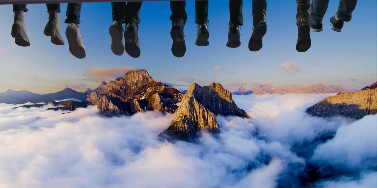 A row of dangling feet in hiking boots above a sea of cloud and sunlit rocky mountain peaks